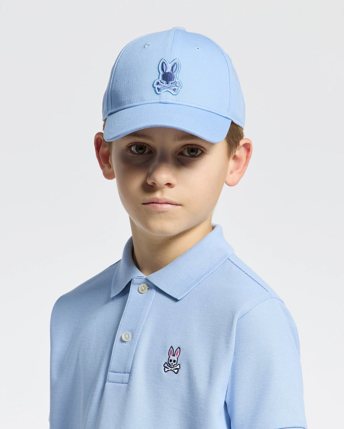 A young boy wears the Psycho Bunny KIDS NEW CASTLE EMBROIDERED BASEBALL CAP - B0A547F200 with an adjustable back strap and a matching light blue polo, both 100% cotton and featuring a cartoon rabbit logo. He stands against a white background, gazing directly at the camera with a neutral expression.