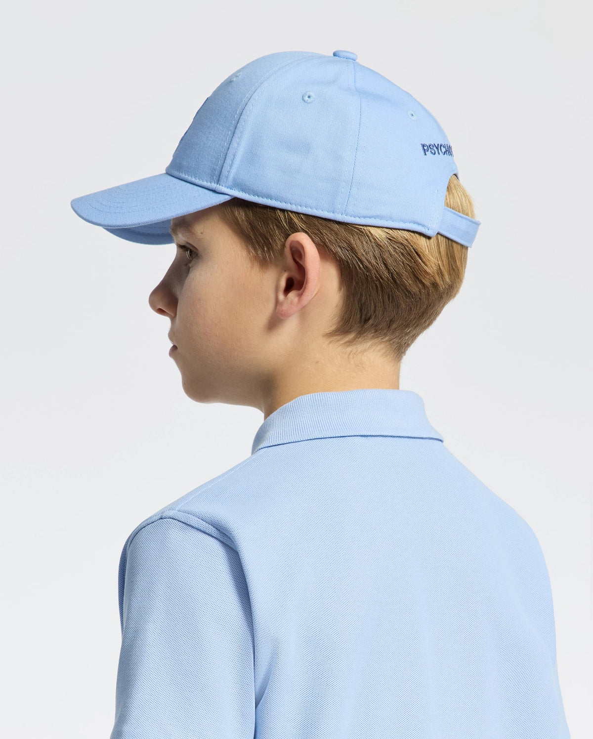 A person with short hair is seen from the back wearing a light blue KIDS NEW CASTLE EMBROIDERED BASEBALL CAP by Psycho Bunny, featuring an adjustable strap, and a matching polo shirt against a plain white background.
