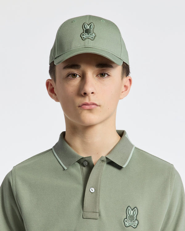A young person wears a sage green cotton twill polo shirt and the KIDS NEW CASTLE BASEBALL CAP - B0A547F200 by Psycho Bunny, adorned with an embroidered bunny logo. The cap features an adjustable back strap. They have a neutral expression against a plain white background.