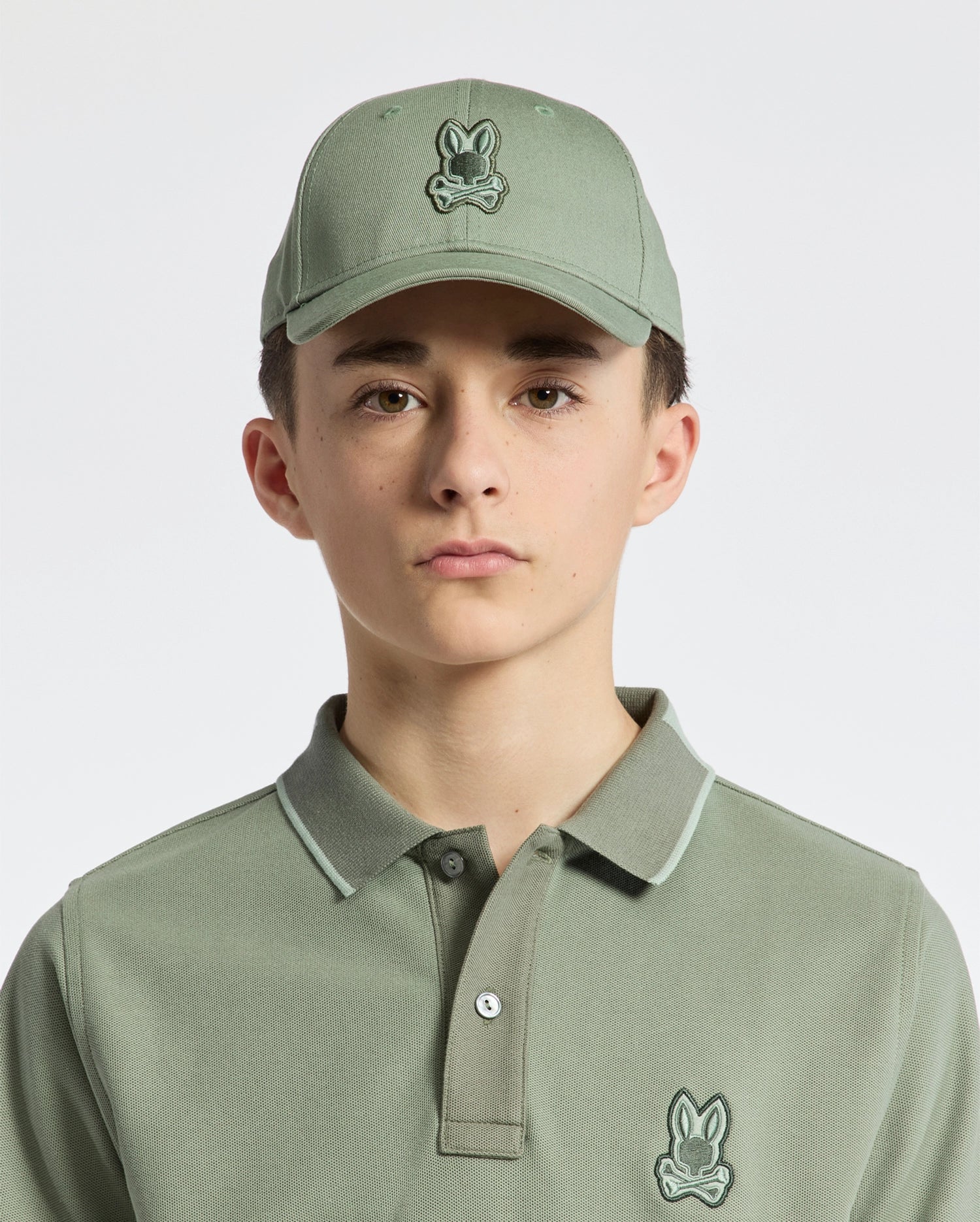 A young person wears a sage green cotton twill polo shirt and the KIDS NEW CASTLE BASEBALL CAP - B0A547F200 by Psycho Bunny, adorned with an embroidered bunny logo. The cap features an adjustable back strap. They have a neutral expression against a plain white background.