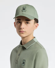 Against a white background, a young person wears a Psycho Bunny KIDS NEW CASTLE BASEBALL CAP in sage green with an adjustable back strap and a polo shirt showcasing an embroidered bunny logo.