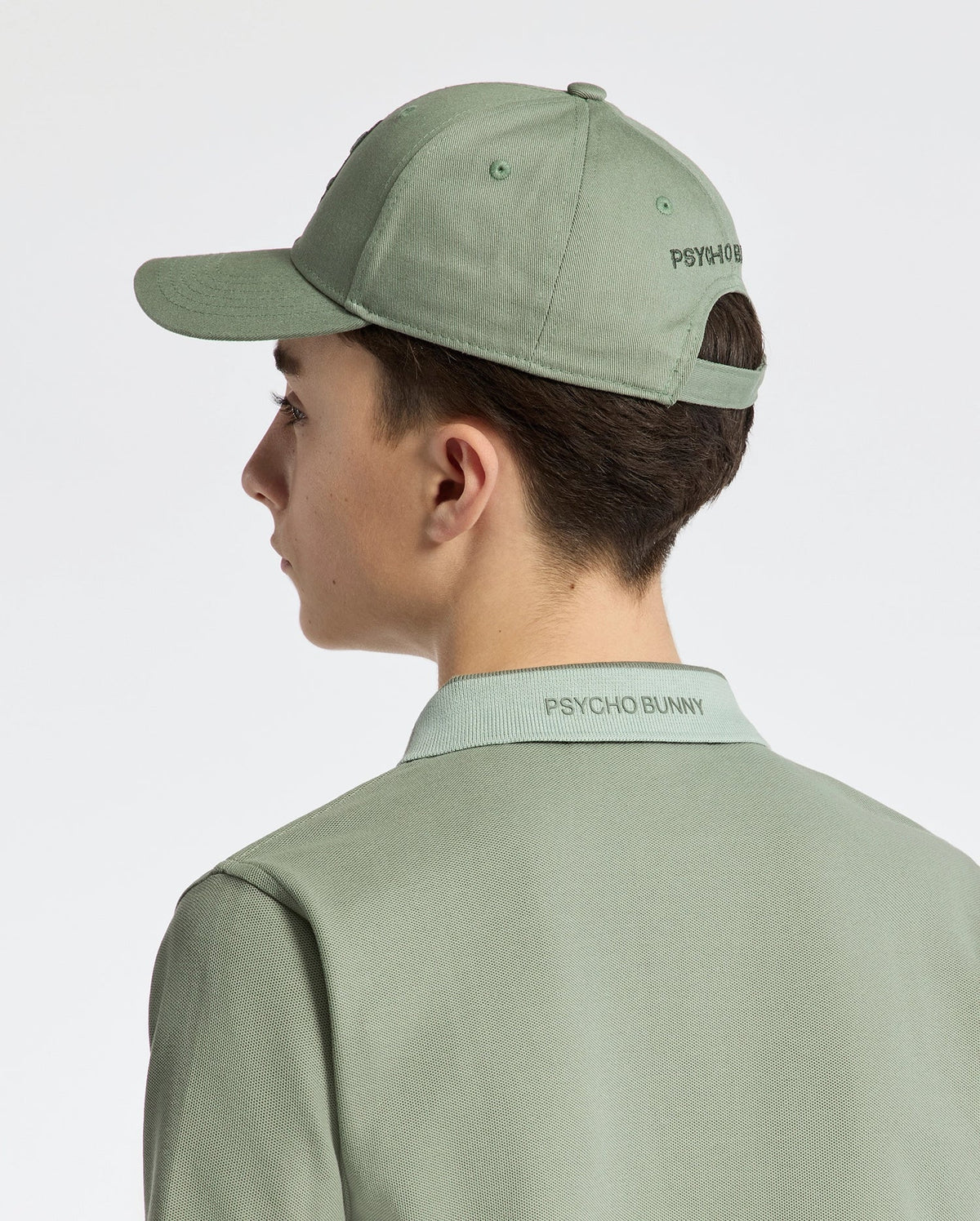 A person in a Psycho Bunny KIDS NEW CASTLE BASEBALL CAP, featuring "Psycho Bunny" text and crafted from cotton twill with an adjustable back strap, wears a matching green polo shirt. They face away against a neutral background.