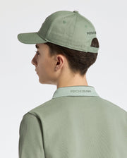 A person in a Psycho Bunny KIDS NEW CASTLE BASEBALL CAP, featuring "Psycho Bunny" text and crafted from cotton twill with an adjustable back strap, wears a matching green polo shirt. They face away against a neutral background.