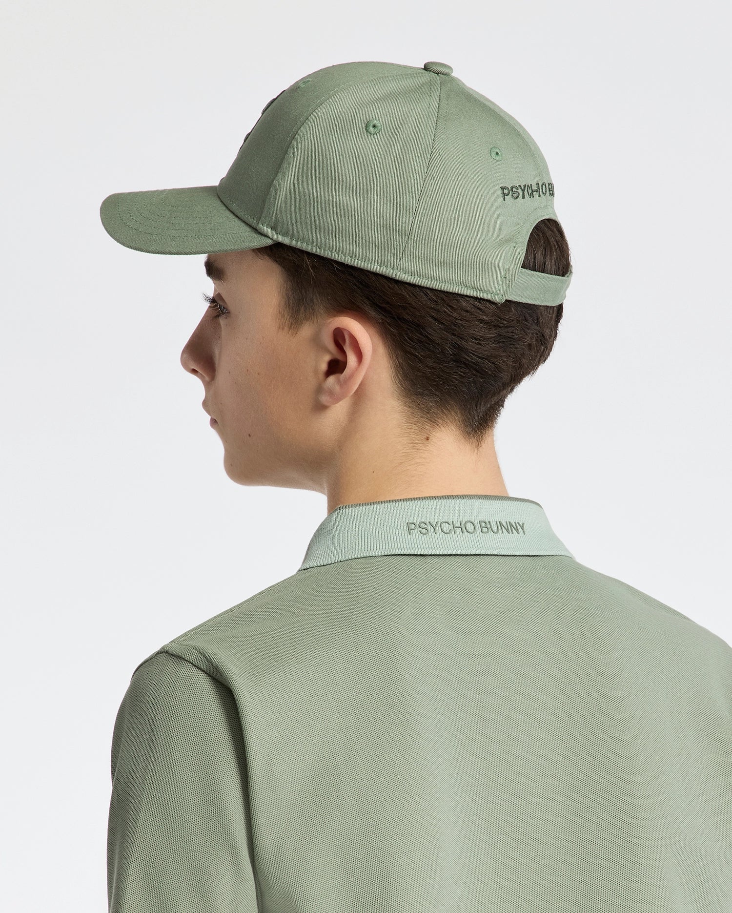 A person in a Psycho Bunny KIDS NEW CASTLE BASEBALL CAP, featuring "Psycho Bunny" text and crafted from cotton twill with an adjustable back strap, wears a matching green polo shirt. They face away against a neutral background.