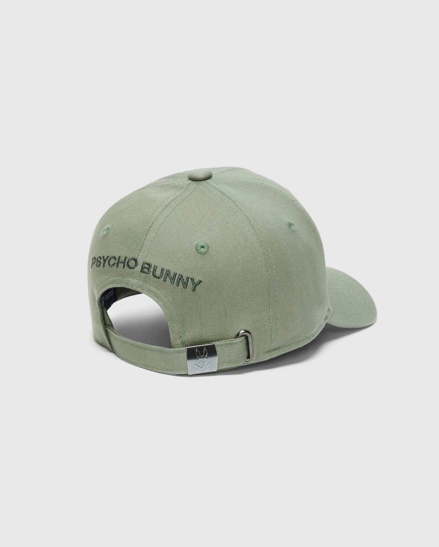 The KIDS NEW CASTLE BASEBALL CAP (B0A547F200) by Psycho Bunny is a sage green cotton twill cap with "PSYCHO BUNNY" embroidered in black on the back. It features an adjustable strap with a metal clasp, and displays a bunny face and crossbones emblem.
