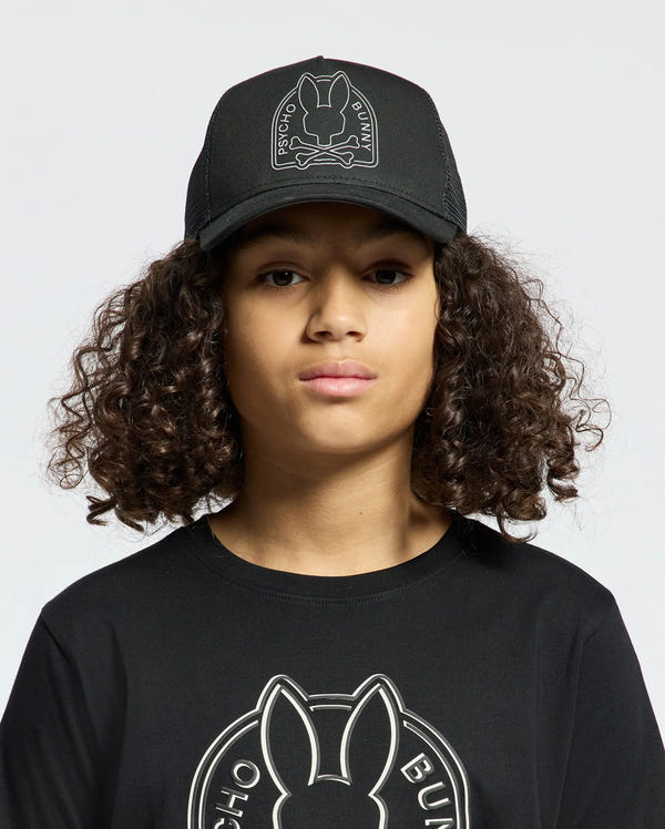 A person with curly hair wears a black Psycho Bunny hat named "Kids Melbourne Graphic Trucker Hat - B0A548F200," along with a matching black shirt. Each piece features an embroidered bunny logo and snap-back straps, against a plain background, while the person faces forward.
