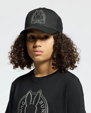 A person with curly hair wears a Psycho Bunny KIDS MELBOURNE GRAPHIC TRUCKER HAT and T-shirt, both with an embroidered bunny icon, against a plain white backdrop.