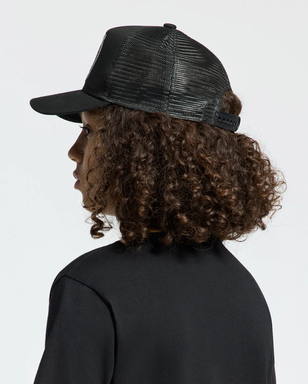 A person with curly brown hair is wearing a black mesh KIDS MELBOURNE GRAPHIC TRUCKER HAT by Psycho Bunny featuring an embroidered icon and a black shirt, gazing to the side against a neutral backdrop.