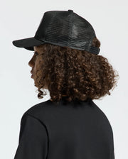 A person with curly brown hair is wearing a black mesh KIDS MELBOURNE GRAPHIC TRUCKER HAT by Psycho Bunny featuring an embroidered icon and a black shirt, gazing to the side against a neutral backdrop.