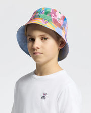 A young person wearing the KIDS MILOS BUCKET REVERSIBLE HAT - B0A550F200 and a plain white shirt with an embroidered Psycho Bunny logo stands against a plain background, looking into the camera.