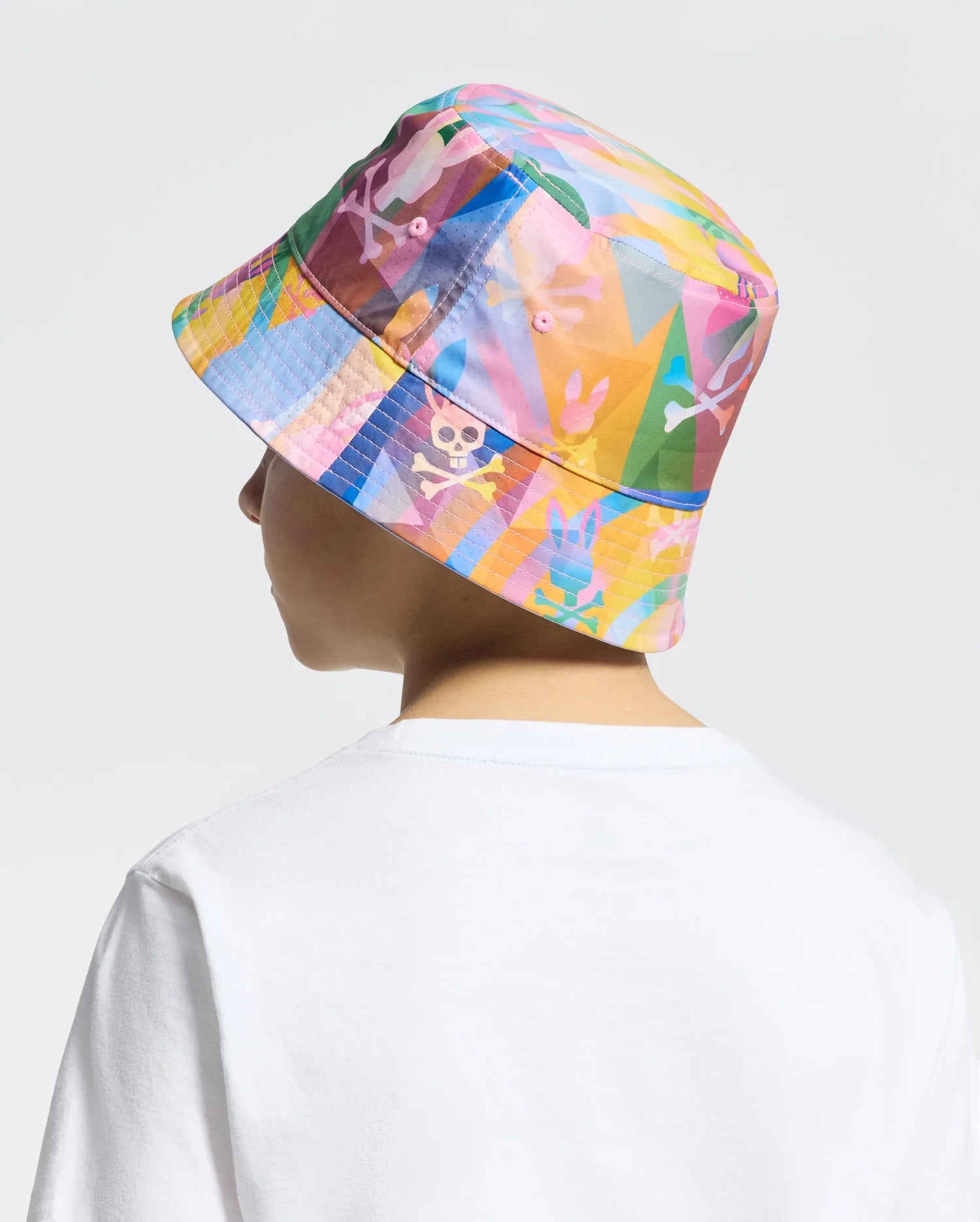 Wearing a colorful KIDS MILOS BUCKET REVERSIBLE HAT (B0A550F200) by Psycho Bunny, adorned with embroidered patterns like skulls and bunny ears, a person in a white shirt stands against a plain background, showcasing the vibrant design.