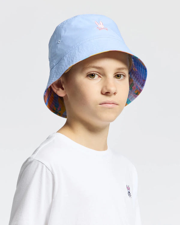A person wearing a light blue KIDS MILOS BUCKET REVERSIBLE HAT by Psycho Bunny, featuring an embroidered pink emblem and a colorful patterned brim, stands against a plain background, paired with a white t-shirt.