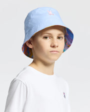 A person wearing a light blue KIDS MILOS BUCKET REVERSIBLE HAT by Psycho Bunny, featuring an embroidered pink emblem and a colorful patterned brim, stands against a plain background, paired with a white t-shirt.