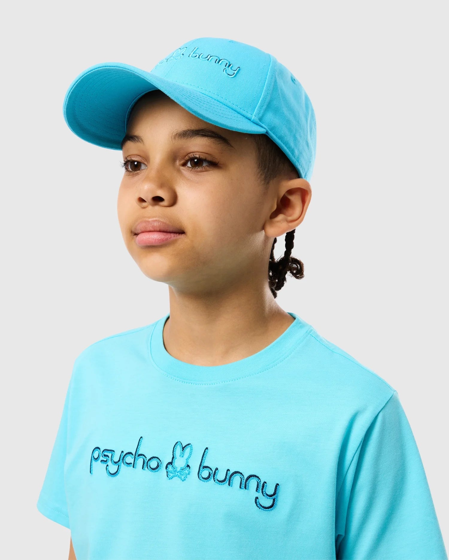 A young person wears the KIDS LOUISE BASEBALL CAP - B0A551C200 from Psycho Bunny, featuring a light blue design and embroidered logo on cotton twill, paired with a matching t-shirt against a plain gray background.