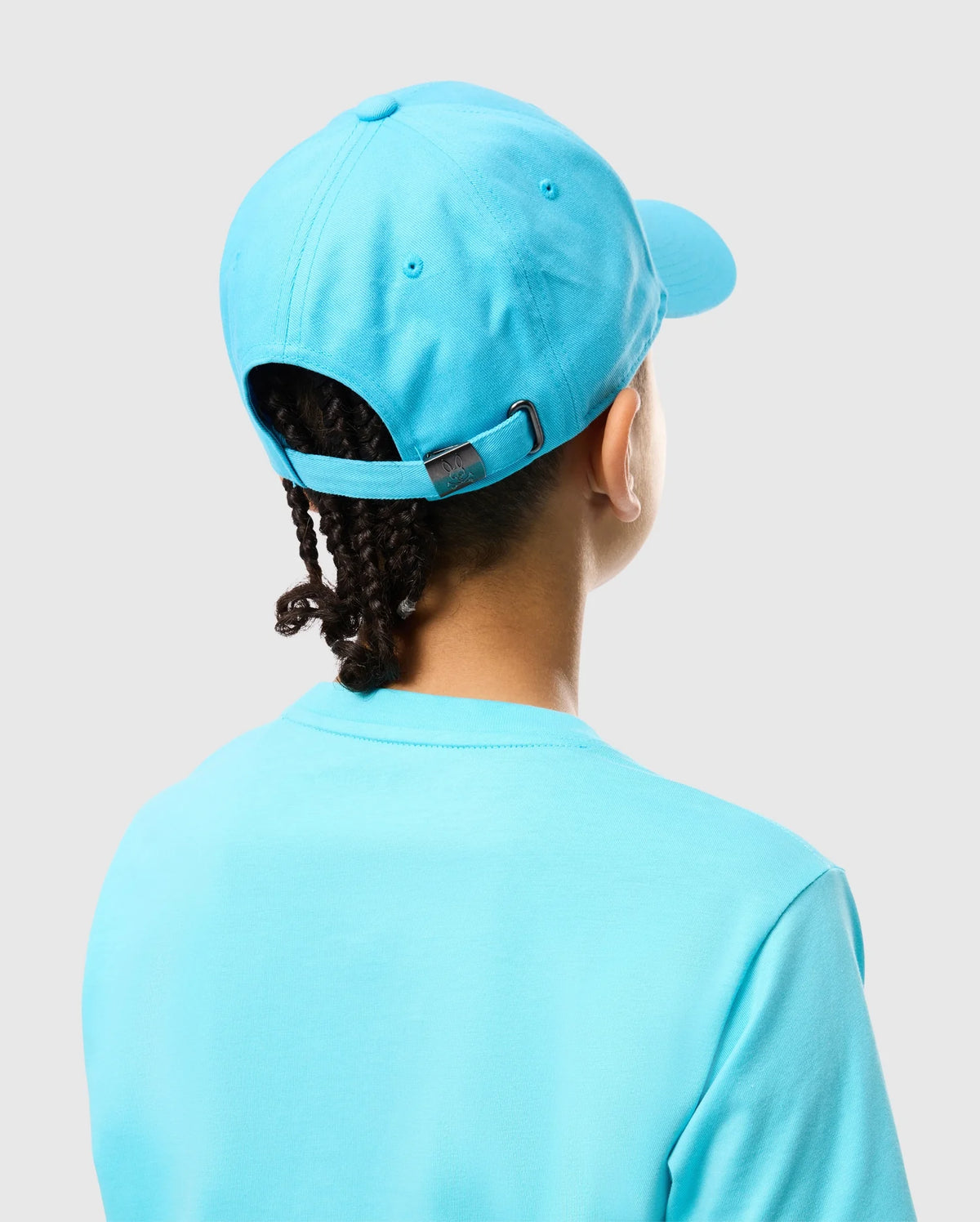 A person in a Psycho Bunny KIDS LOUISE BASEBALL CAP - B0A551C200, bright blue cotton twill with an embroidered logo and metal buckle, faces away, showing braided hair peeking out. They wear a matching t-shirt against a light gray background.