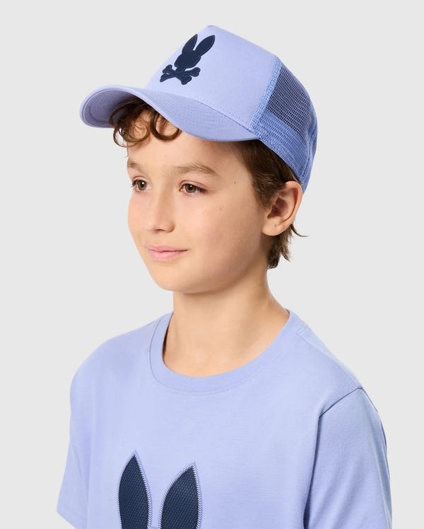 A young boy with curly hair smiles while wearing a light blue Psycho Bunny KIDS RIVIERA TRUCKER CAP, complemented by a matching T-shirt. Both items are adorned with a dark rabbit and crossbones logo, while the snapback fastening of the cap lends a hint of 2000s nostalgia against the plain gray background.