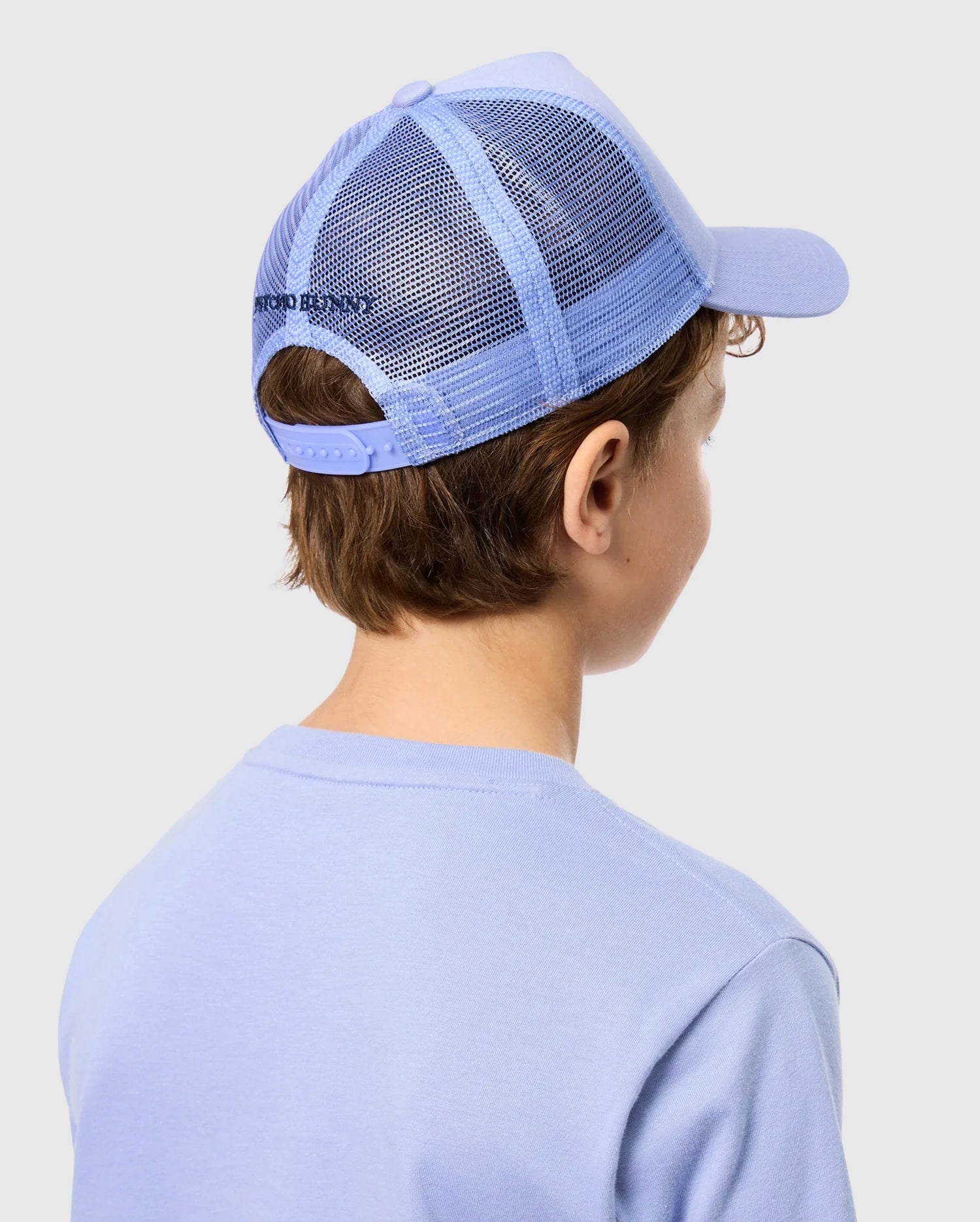 A person with brown hair wears a blue Psycho Bunny KIDS RIVIERA TRUCKER CAP - B0A667C200 and a matching shirt, channeling 2000s nostalgia. They are turned away, showcasing the cap's adjustable snapback fastening against a simple light gray backdrop.