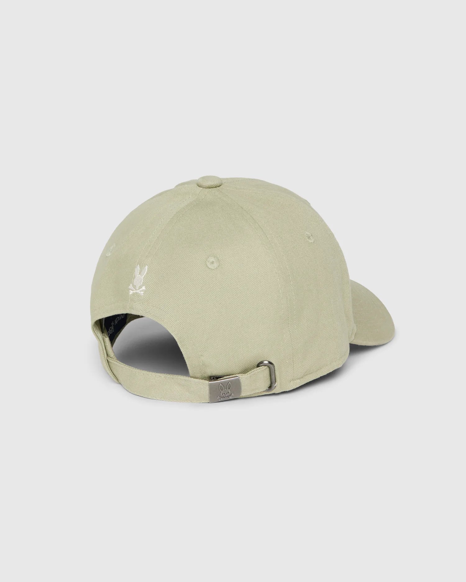 A light beige cotton twill baseball cap from Psycho Bunny, named the Kids Kayden Baseball Cap - B0A677C200, is viewed from the back. The cap features a metal buckle for size adjustment and an embroidered Bunny logo above the opening. The fabric has a smooth texture, and the cap is displayed against a plain, neutral background.