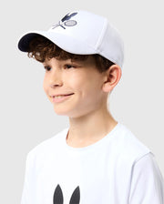A young boy wears a Psycho Bunny KIDS COURTSIDE SPORT CAP - B0A685C200, featuring crossed tennis rackets. His white t-shirt complements his curly brown hair as he smiles against a plain light gray backdrop.