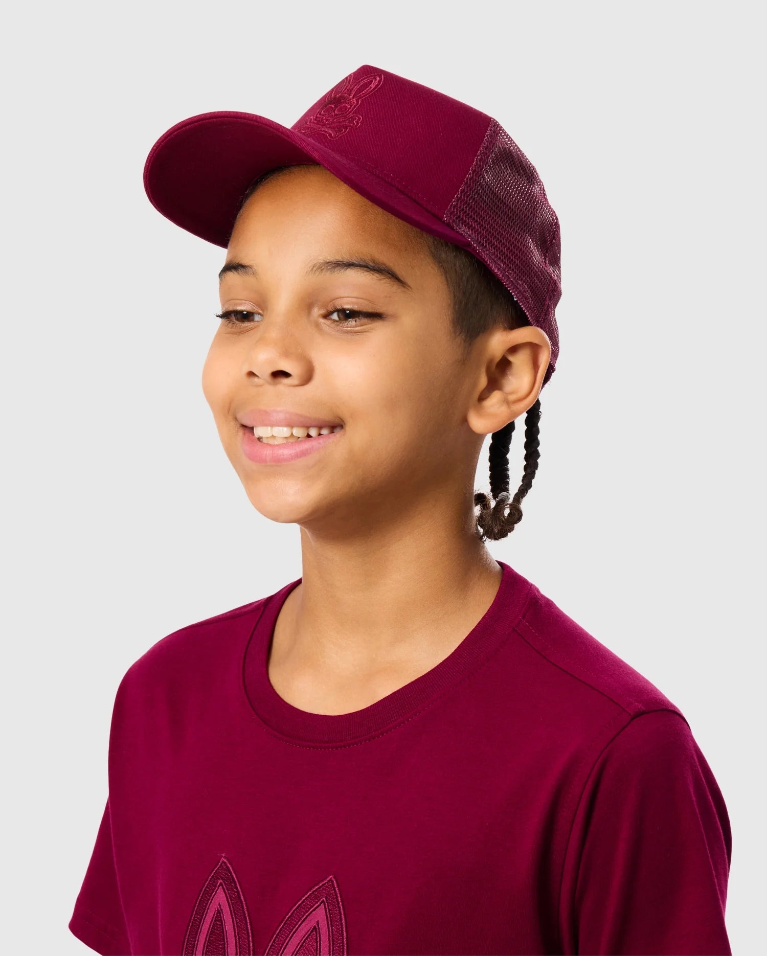 A young person smiles wearing a maroon KIDS MALTON TRUCKER CAP (B0A797D200) and matching T-shirt by Psycho Bunny, featuring mesh detailing on the back, set against a light gray background.