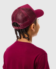 The image features a person from behind wearing the Psycho Bunny burgundy KIDS MALTON TRUCKER CAP - B0A797D200 and matching t-shirt. They have braided hair and stand against a light gray background, showcasing an accessory that blends an active lifestyle with effortless style.