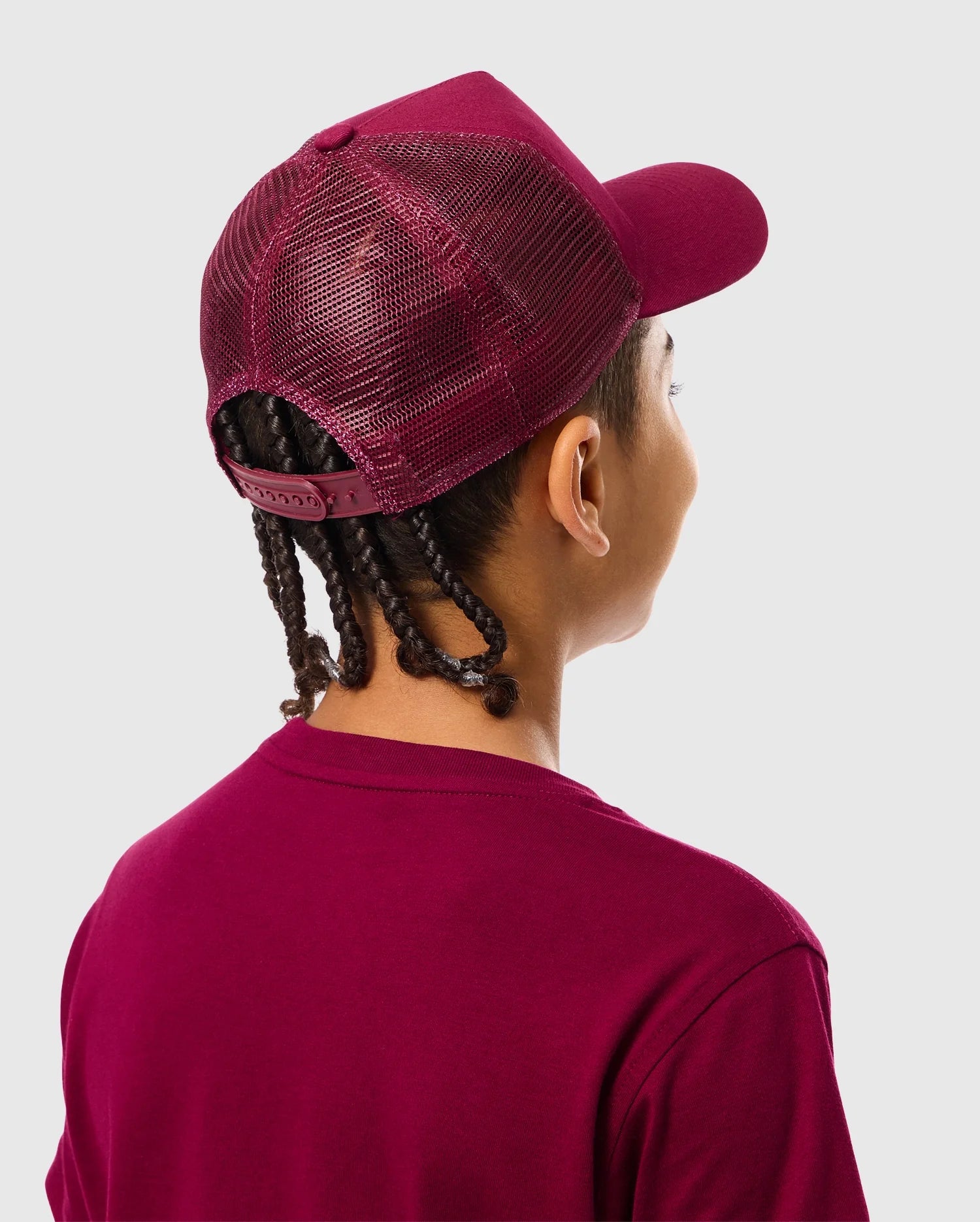 The image features a person from behind wearing the Psycho Bunny burgundy KIDS MALTON TRUCKER CAP - B0A797D200 and matching t-shirt. They have braided hair and stand against a light gray background, showcasing an accessory that blends an active lifestyle with effortless style.