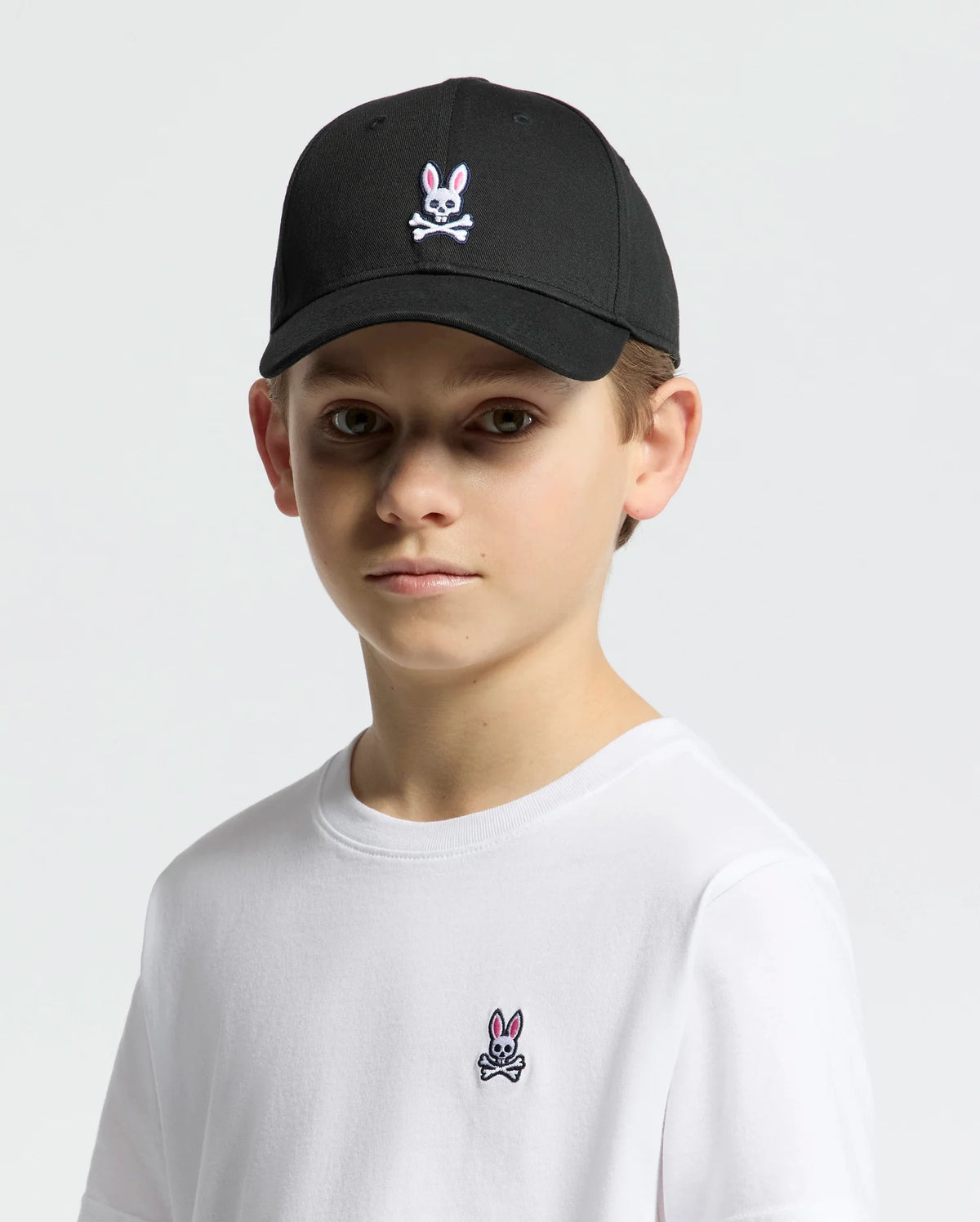 A child wears a Psycho Bunny KIDS CLASSIC BASEBALL CAP - B0A816A1HT in black featuring an embroidered bunny and crossbones, perfectly paired with a white t-shirt displaying the same logo. The cap's adjustable fit ensures style and comfort against a plain white background.