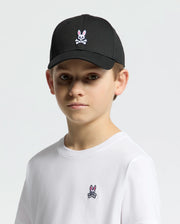 A child wears a Psycho Bunny KIDS CLASSIC BASEBALL CAP - B0A816A1HT in black featuring an embroidered bunny and crossbones, perfectly paired with a white t-shirt displaying the same logo. The cap's adjustable fit ensures style and comfort against a plain white background.
