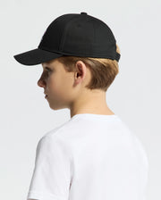 A young boy in a white T-shirt, wearing a Psycho Bunny Kids Classic Baseball Cap (B0A816A1HT), is shown in profile against a plain background. The adjustable cap, slightly tilted forward, obscures part of his face while his blonde hair peeks out beneath the embroidered Bunny logo at the back.