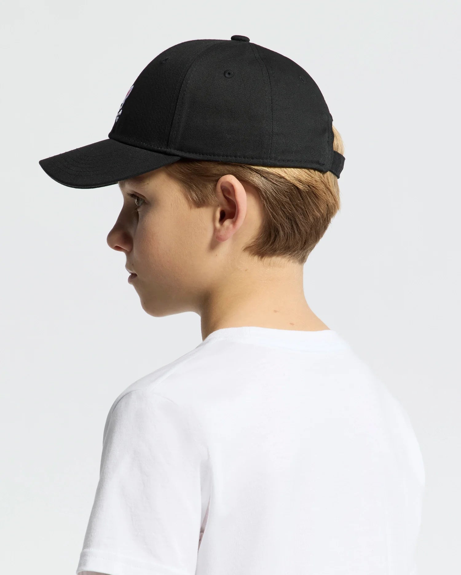 A young boy in a white T-shirt, wearing a Psycho Bunny Kids Classic Baseball Cap (B0A816A1HT), is shown in profile against a plain background. The adjustable cap, slightly tilted forward, obscures part of his face while his blonde hair peeks out beneath the embroidered Bunny logo at the back.
