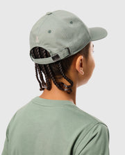 A person in a sporty, sage green KIDS ZACK BASEBALL CAP - B0A886D200 from Psycho Bunny and a matching t-shirt is shown from behind. The woven cotton cap, featuring a small embroidered logo, complements their braided hair against a plain light gray background.