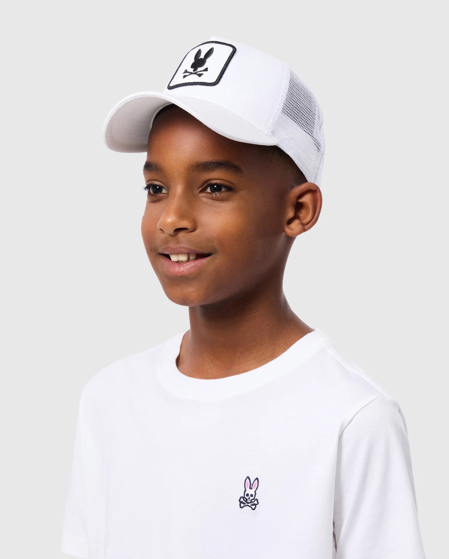 A boy wears the Psycho Bunny Kids Bunny Patch Trucker Cap, showcasing a black and white animal skull emblem. His crisp white T-shirt with a subtle bunny skull logo complements the cap, perfectly offset against a plain gray background, highlighting his cool style.