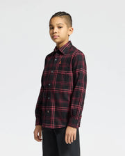 A young person with braided hair confidently gazes at the camera, wearing a Psycho Bunny KIDS HAYES PLAID SHIRT in red and black with black pants, set against a plain light gray background.