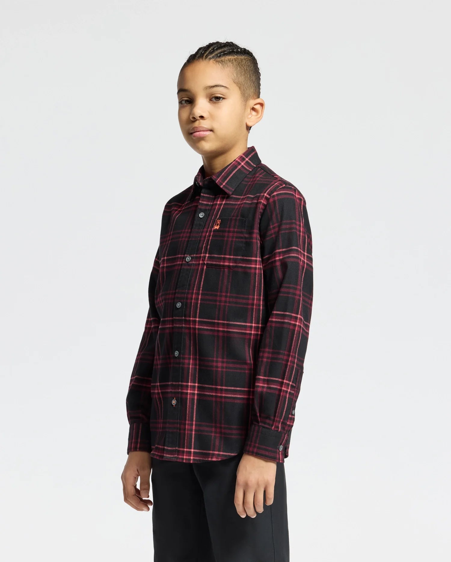 A young person with braided hair confidently gazes at the camera, wearing a Psycho Bunny KIDS HAYES PLAID SHIRT in red and black with black pants, set against a plain light gray background.