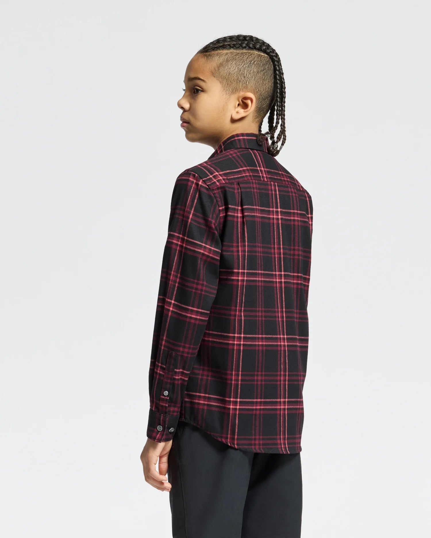 A young person with braided hair is slightly turned away from the camera, wearing a Psycho Bunny KIDS HAYES PLAID SHIRT - B0C842D200 in black and red Pima cotton, against a plain white background.