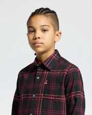 A young person with braided hair wears the Psycho Bunny KIDS HAYES PLAID SHIRT B0C842D200 in black and red, featuring a small embroidered logo on the chest, against a plain background. They look directly at the camera with a neutral expression.