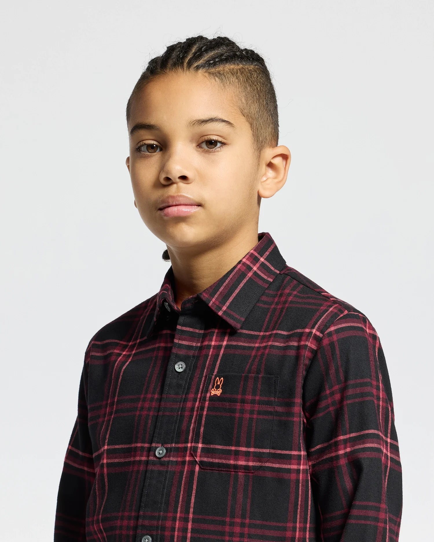 A young person with braided hair wears the Psycho Bunny KIDS HAYES PLAID SHIRT B0C842D200 in black and red, featuring a small embroidered logo on the chest, against a plain background. They look directly at the camera with a neutral expression.