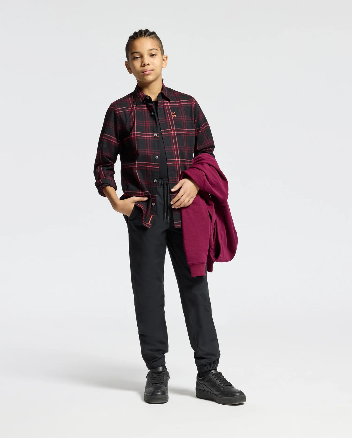 A person with braided hair stands confidently, wearing a KIDS HAYES PLAID SHIRT in black from Psycho Bunny over a black top and pants. Holding a red jacket in their left hand and donning black boots, they exude style against the crisp white background that highlights their sleek ensemble's bold colors.
