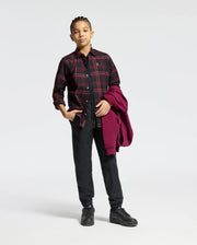 A person with braided hair stands confidently, wearing a KIDS HAYES PLAID SHIRT in black from Psycho Bunny over a black top and pants. Holding a red jacket in their left hand and donning black boots, they exude style against the crisp white background that highlights their sleek ensemble's bold colors.