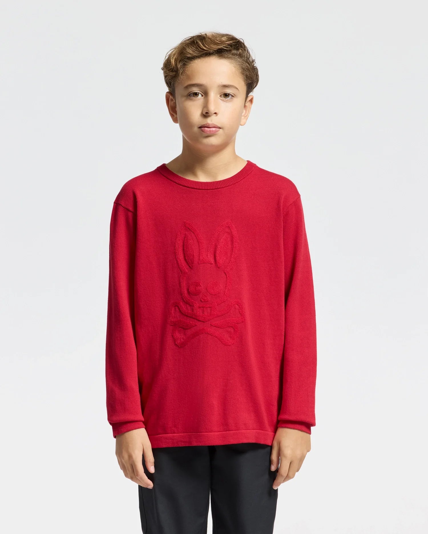 A young boy with short brown hair wears a red Psycho Bunny Kids Coory Chainstitch Cotton Sweater, featuring chainstitch embroidery of a stylized bunny and crossbones on the front. He stands against a plain white background, wearing black pants.