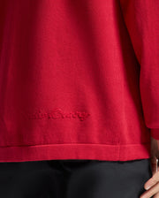 A person wearing a red KIDS COORY CHAINSTITCH COTTON SWEATER by Psycho Bunny, featuring "Gucci Camp" subtly chainstitched on the lower back. Made from extra fine cotton, it pairs perfectly with black pants. Only the lower back and part of the arm are visible.