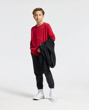 A young person stands confidently in Psycho Bunny's KIDS COORY CHAINSTITCH COTTON SWEATER, paired with black pants and white high-top sneakers, holding a black jacket over their arm against a plain white background.