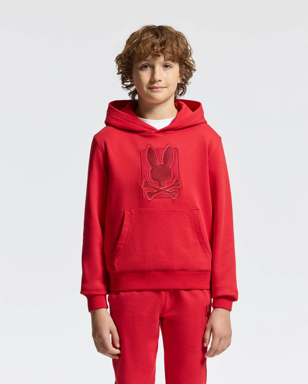 A young person with curly hair is wearing the KIDS PIERCE ESSENTIAL FRENCH TERRY HOODIE from Psycho Bunny, along with matching joggers. The red pullover sweatshirt showcases an embossed design of a bunny with crossed bones beneath it. The plain and well-lit background emphasizes the vibrant kids' cotton ensemble.