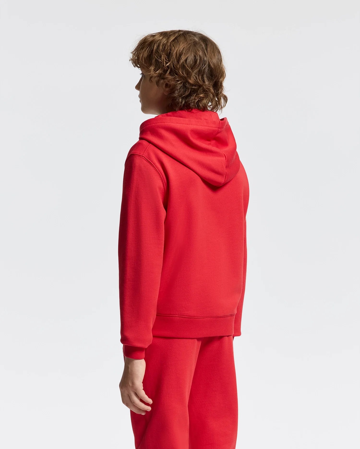 A person with curly hair is seen from the back wearing a red KIDS PIERCE ESSENTIAL FRENCH TERRY HOODIE by Psycho Bunny and matching red pants against a plain white background.