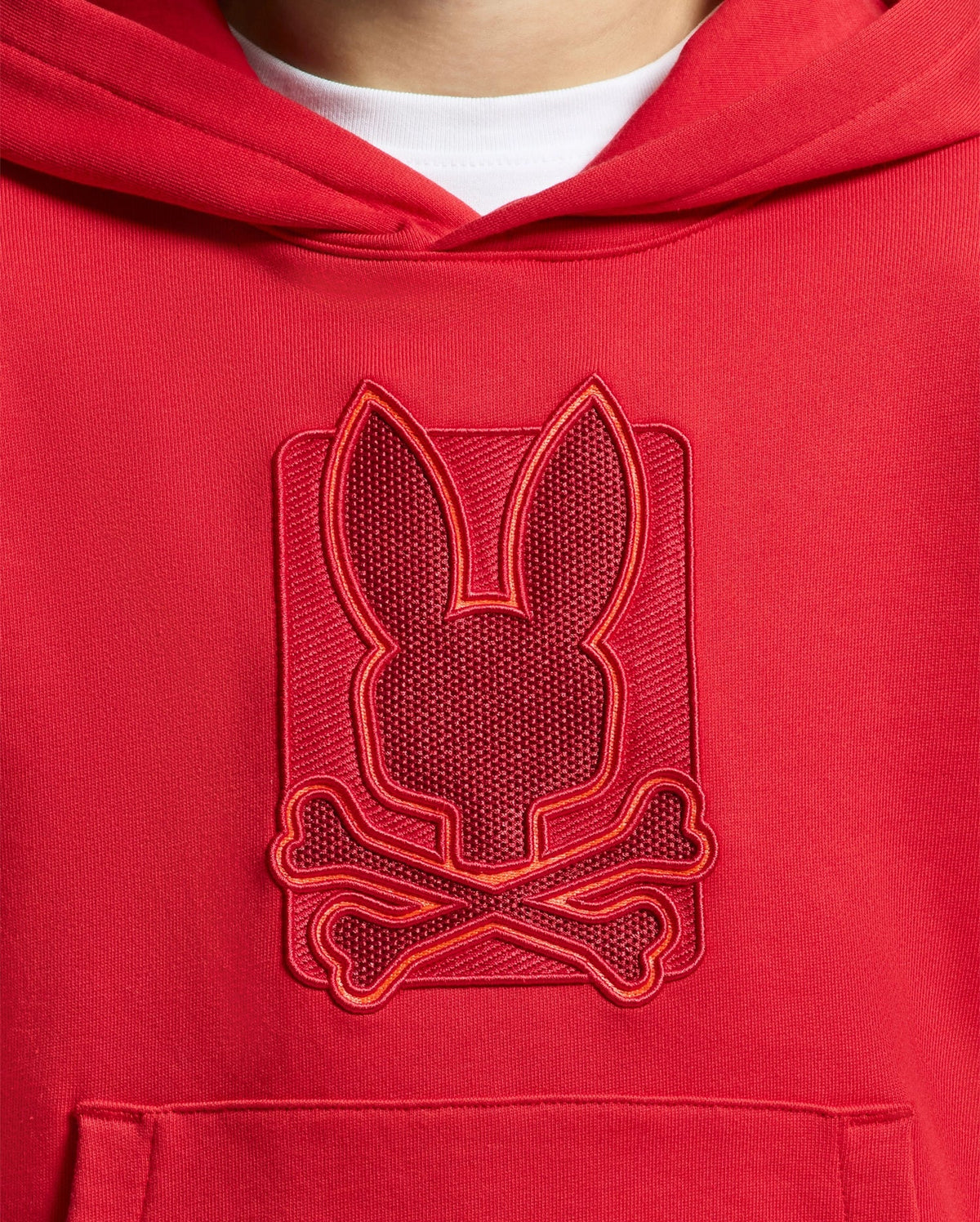 A close-up of the KIDS PIERCE ESSENTIAL FRENCH TERRY HOODIE from Psycho Bunny highlights a textured emblem featuring a stylized bunny head above crossed bones on the chest. The intricately embroidered design uses red and orange threads, making this pullover sweatshirt stand out brilliantly.