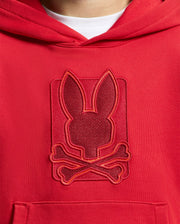 A close-up of the KIDS PIERCE ESSENTIAL FRENCH TERRY HOODIE from Psycho Bunny highlights a textured emblem featuring a stylized bunny head above crossed bones on the chest. The intricately embroidered design uses red and orange threads, making this pullover sweatshirt stand out brilliantly.