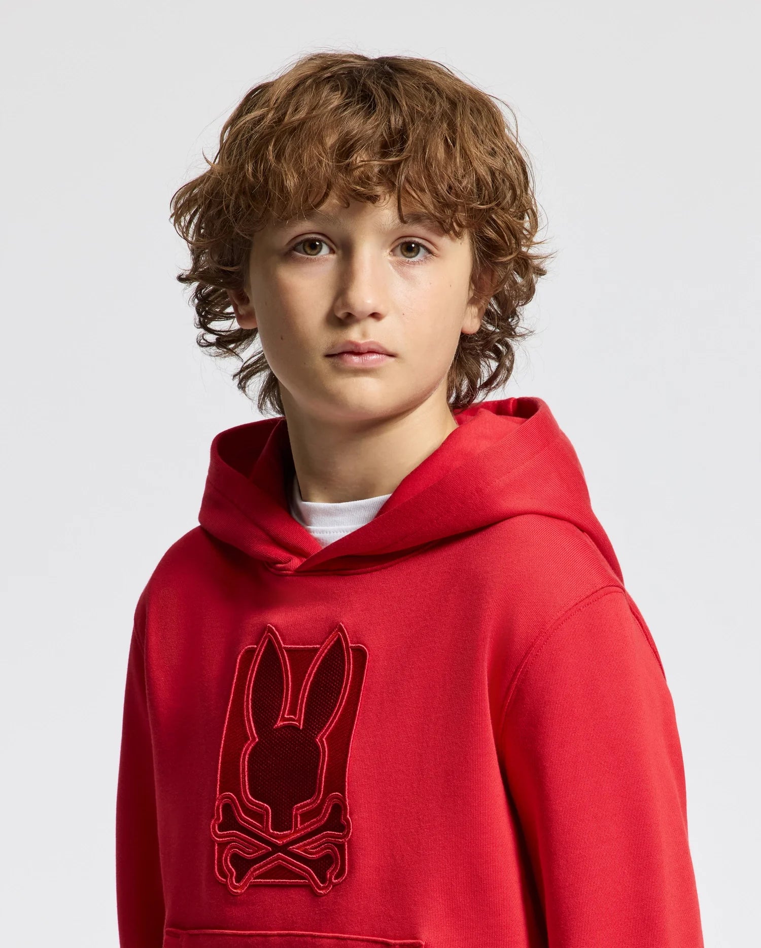 A young person with curly hair is wearing a vibrant Psycho Bunny KIDS PIERCE ESSENTIAL FRENCH TERRY HOODIE (B0H135E200), featuring a rabbit and crossbones design on the front, standing against a plain white background.