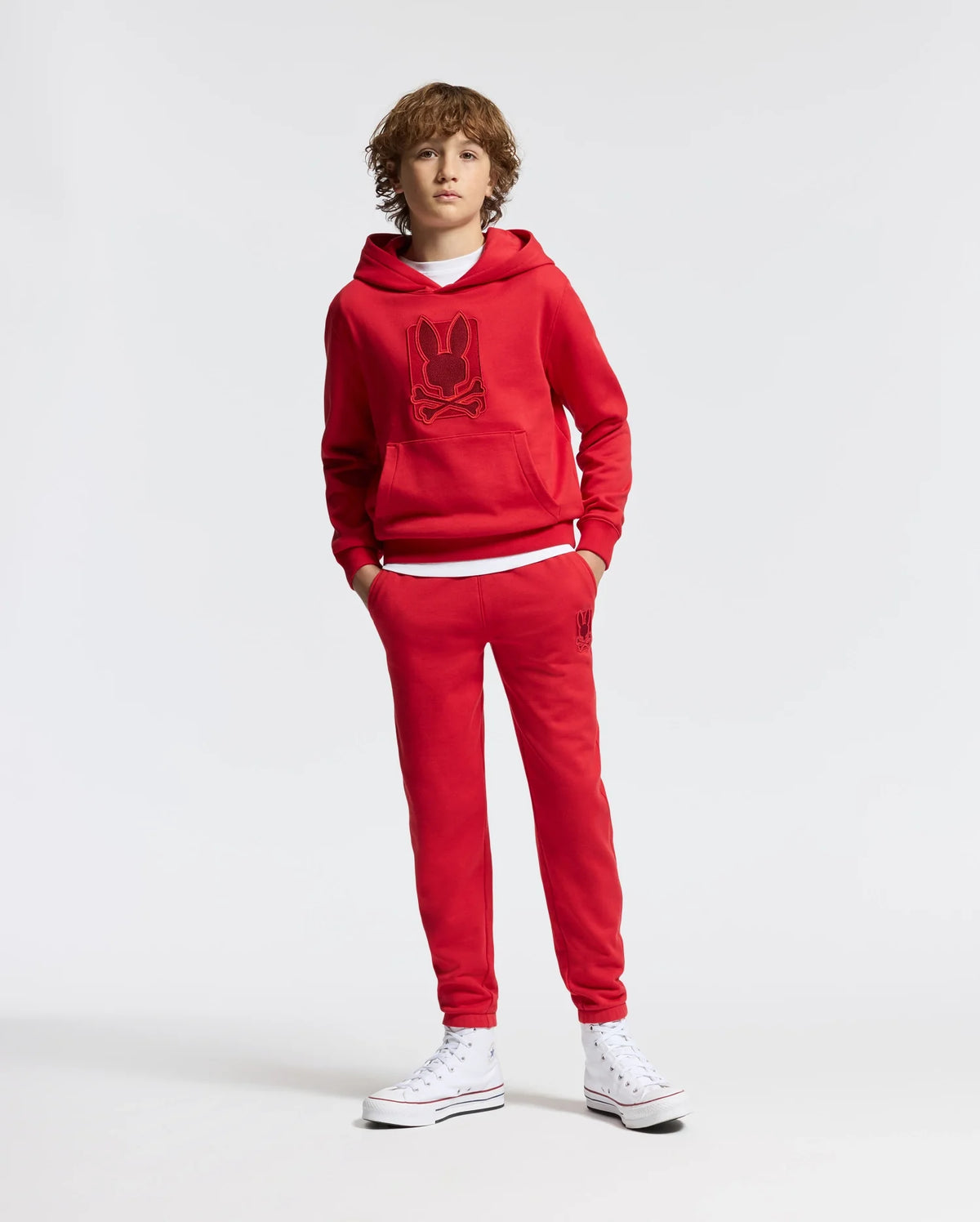 A person with curly hair is dressed in a red KIDS PIERCE ESSENTIAL FRENCH TERRY HOODIE by Psycho Bunny, paired with matching red sweatpants featuring a minimalistic graphic. They stand upright against a plain white background, wearing high-top white sneakers.
