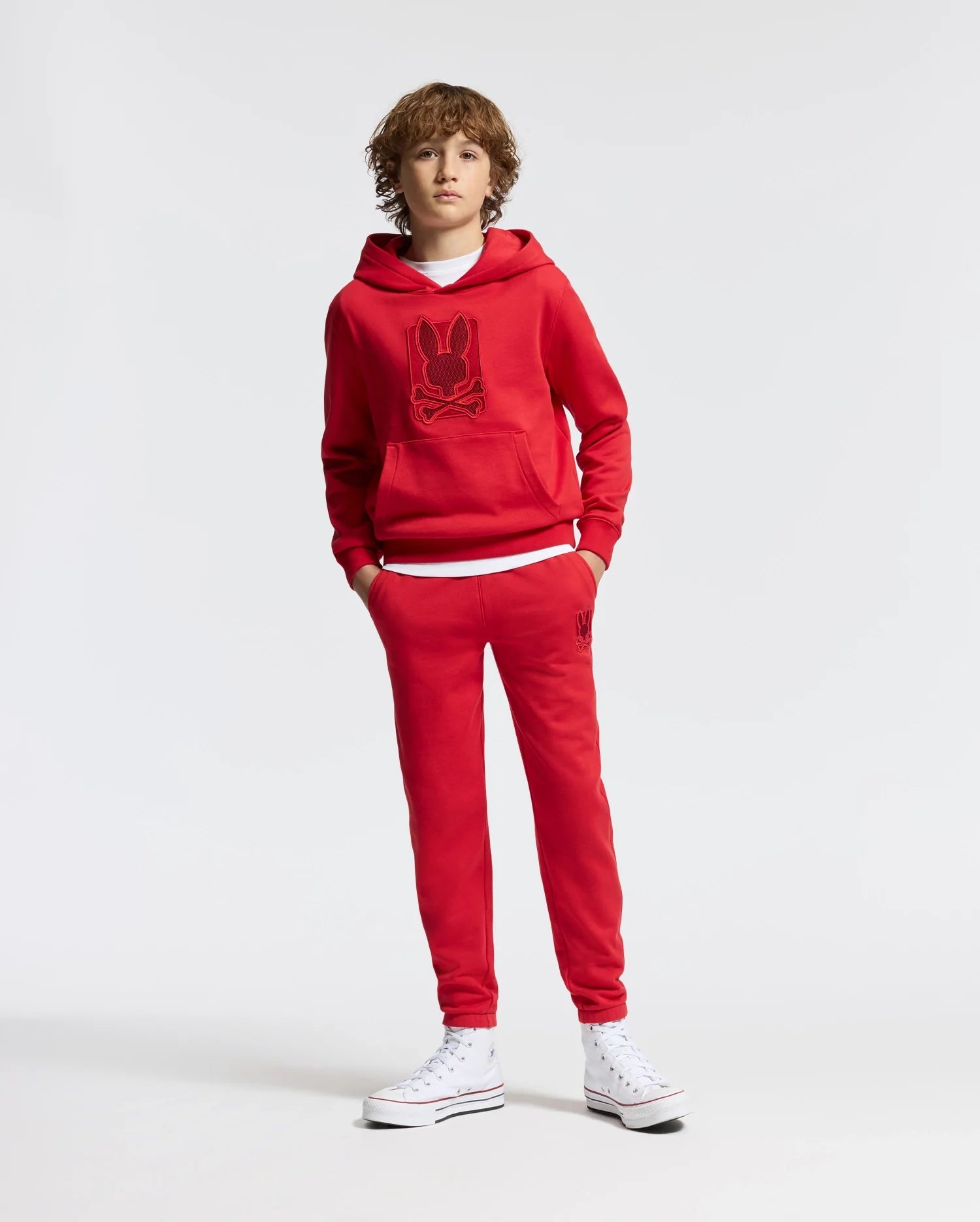 A person with curly hair is dressed in a red KIDS PIERCE ESSENTIAL FRENCH TERRY HOODIE by Psycho Bunny, paired with matching red sweatpants featuring a minimalistic graphic. They stand upright against a plain white background, wearing high-top white sneakers.