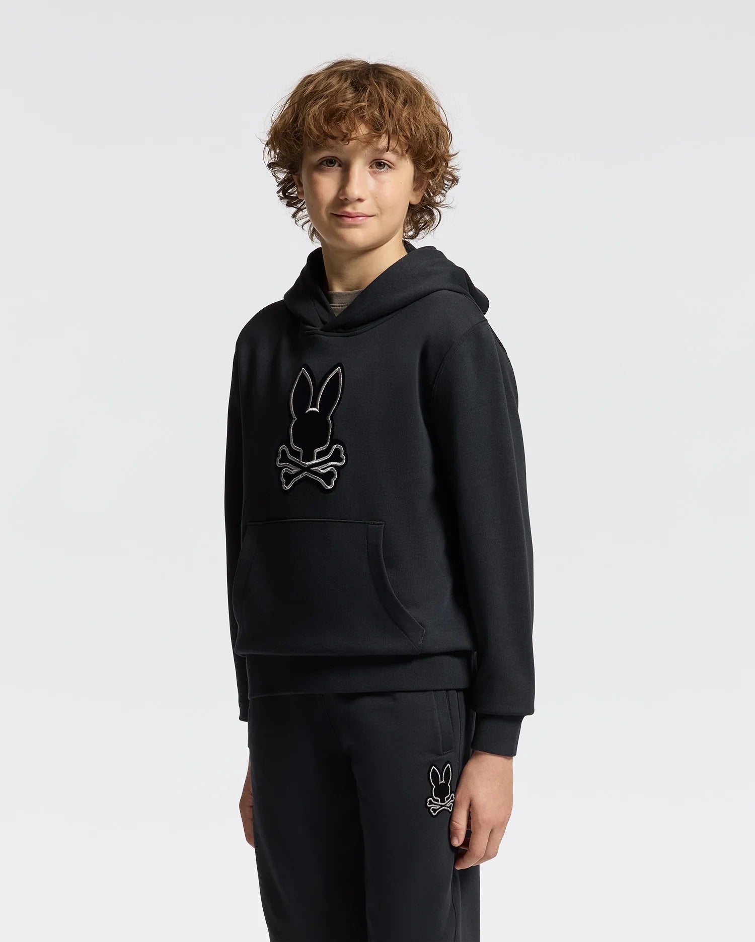 A young person with curly hair is wearing the Psycho Bunny KIDS ALEXANDER HOODIE - B0H154E200, a black hoodie featuring a stylized bunny logo, along with sweatpants. This versatile pullover offers a comfortable relaxed fit against a plain white background.